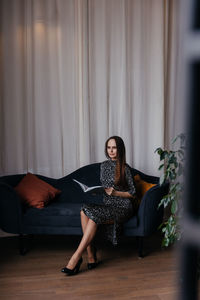 A woman in a dress with polka dots and classic black shoes sits on a dark blue sofa 