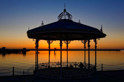 Silhouette built structure by sea against orange sky