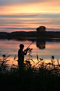 Silhouette man fishing by lake against sky during sunset