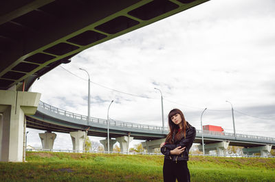 Low angle portrait of confident woman standing on field under bridge