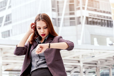 Young businesswoman checking time while standing against buildings in city