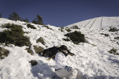 Woman with dog relaxing on snow covered landscape against clear blue sky