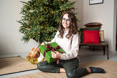 A cute girl with glasses is sitting near a christmas tree and holding a green box with a gift