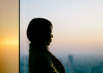 Woman standing by window during sunset