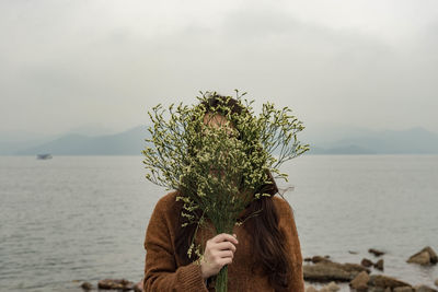 Woman covering face with flowers by sea against sky