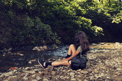 Side view of girl sitting on stones by stream in forest