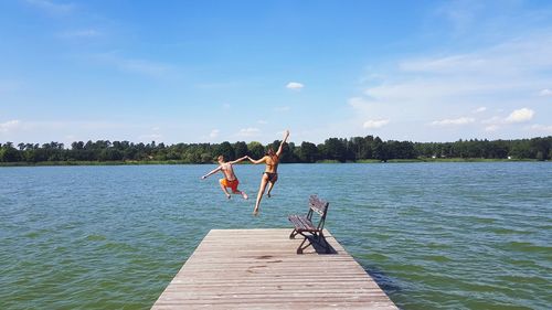 Escaping the heat - rear view of teenage boy and girl jumping on pier over lake against sky