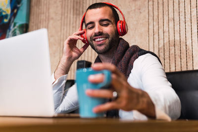 Low angle of cheerful man with cup of coffee smiling and touching wireless headphones while sitting at table with laptop and listening to music during remote work from cafeteria