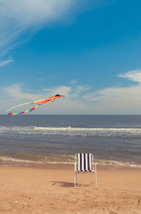 Kite flying over empty chair at beach against sky