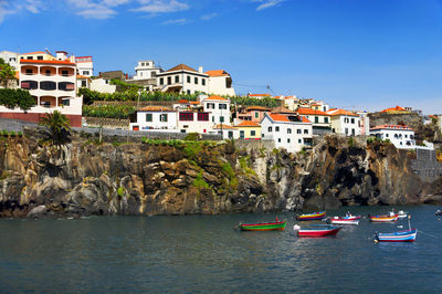 Boats moored in sea by houses on cliff against sky