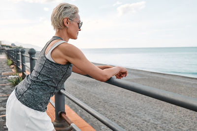 Fit woman relaxes leaning on handrail along seaside promenade after running on a road by the sea. 