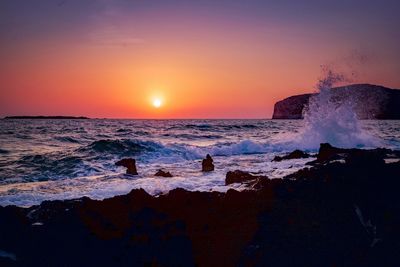 Waves splashing on rocks at shore against sky during sunset