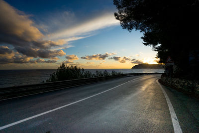 Road by trees against sky during sunset