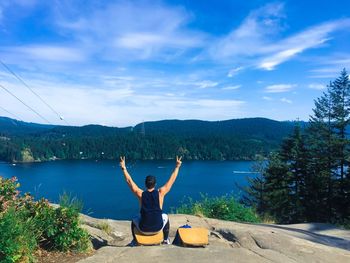 Rear view of man gesturing while sitting at lakeshore against sky