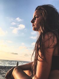 Thoughtful young woman sitting at beach against sky