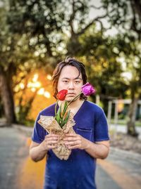 Portrait of young asian man holding red and purple tulips against trees and setting sun.