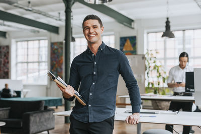 Portrait of happy businessman holding files while leaning on desk at office