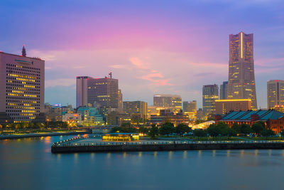 Illuminated buildings by river against sky during sunset