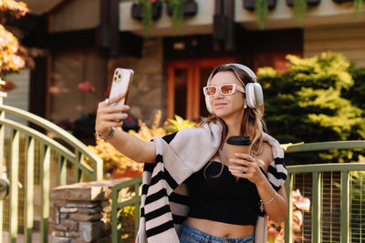 Woman influencer blogger with glasses and headphones looks into a mobile phone on street in autumn