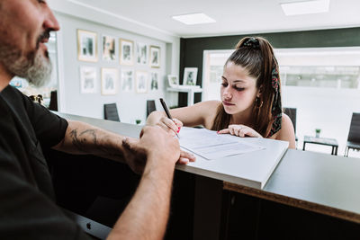 Young female standing at counter in tattoo salon and signing document before procedure