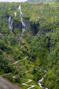 A saterfall in norway with a hiking trail winds sideways up the mountain through dense forest 