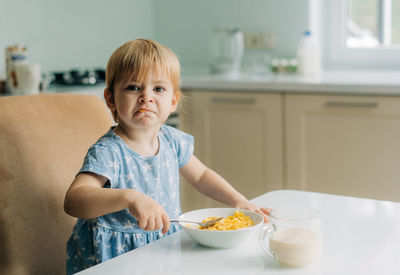 Little toddler girl eats corn flakes with milk in the kitchen in the morning.