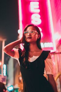 Beautiful young woman standing against illuminated sign in city at night