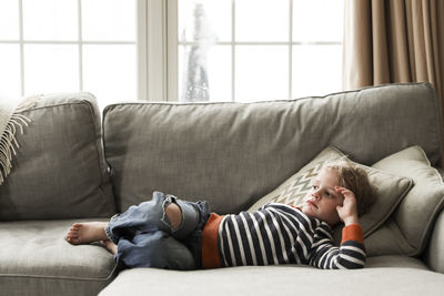 Thoughtful boy lying on sofa against windows at home