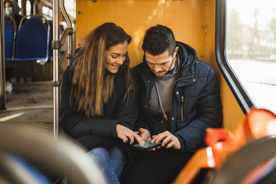 Heterosexual couple using smart phone while sitting in tram during pandemic