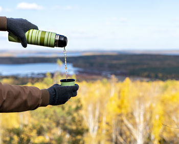 Male hands in warm gloves pour hot tea from thermos against autumn view of lake, yellow forest, fall