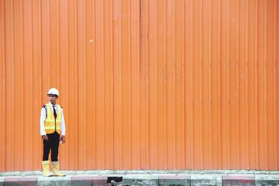 Manual worker wearing reflective clothing and hardhat standing against orange corrugated iron