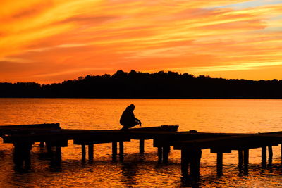 Silhouette man sitting by lake against orange sky