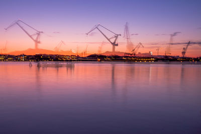 Cranes at harbor against sky during sunset