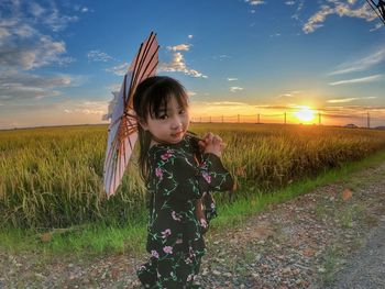 Portrait of girl holding umbrella while standing at farm against sky during sunset