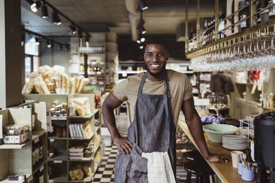 Portrait of smiling male owner with hand on hip standing in delicatessen shop