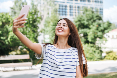 Portrait of young woman student with long hair taking selfie in city park