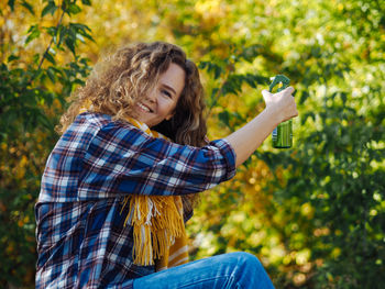 Young curly woman traveller using hair spray in autumn forest