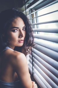 Portrait of beautiful young woman standing by blinds at home