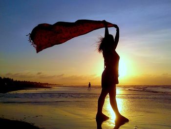 Full length of woman holding scarf while standing at beach during sunset