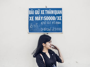 Young woman looking away while standing against wall with signboard