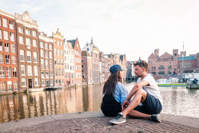 Friends talking while sitting by canal in city against sky