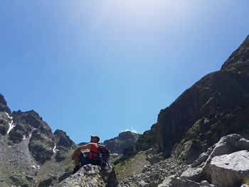 Low angle view of hiker sitting on rocks against clear blue sky during sunny day