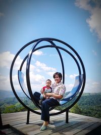 Full length of smiling mother with son sitting against sky