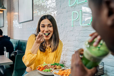 Content female enjoying tasty dish while interacting with crop unrecognizable ethnic partner drinking smoothie in restaurant