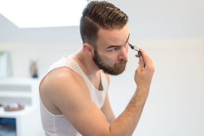Side view of young man shaving beard at home