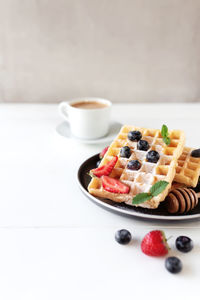Stack of waffles on a plate on white table with blueberry, chopped strawberry, mint, cup of coffee