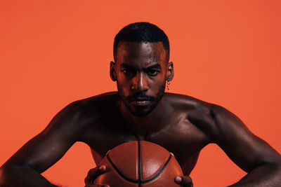 Portrait of shirtless man holding basketball against orange background