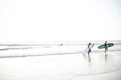 Men with surfboards walking on seashore against clear sky