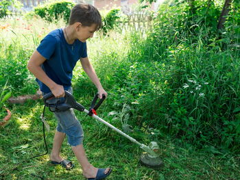 A boy, 11 years old, mows the grass with an electric scythe on the lawn in the yard in summer day.