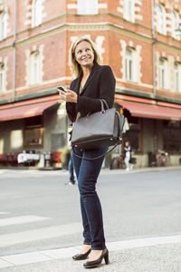 Full length of smiling businesswoman looking away while using mobile phone on city street